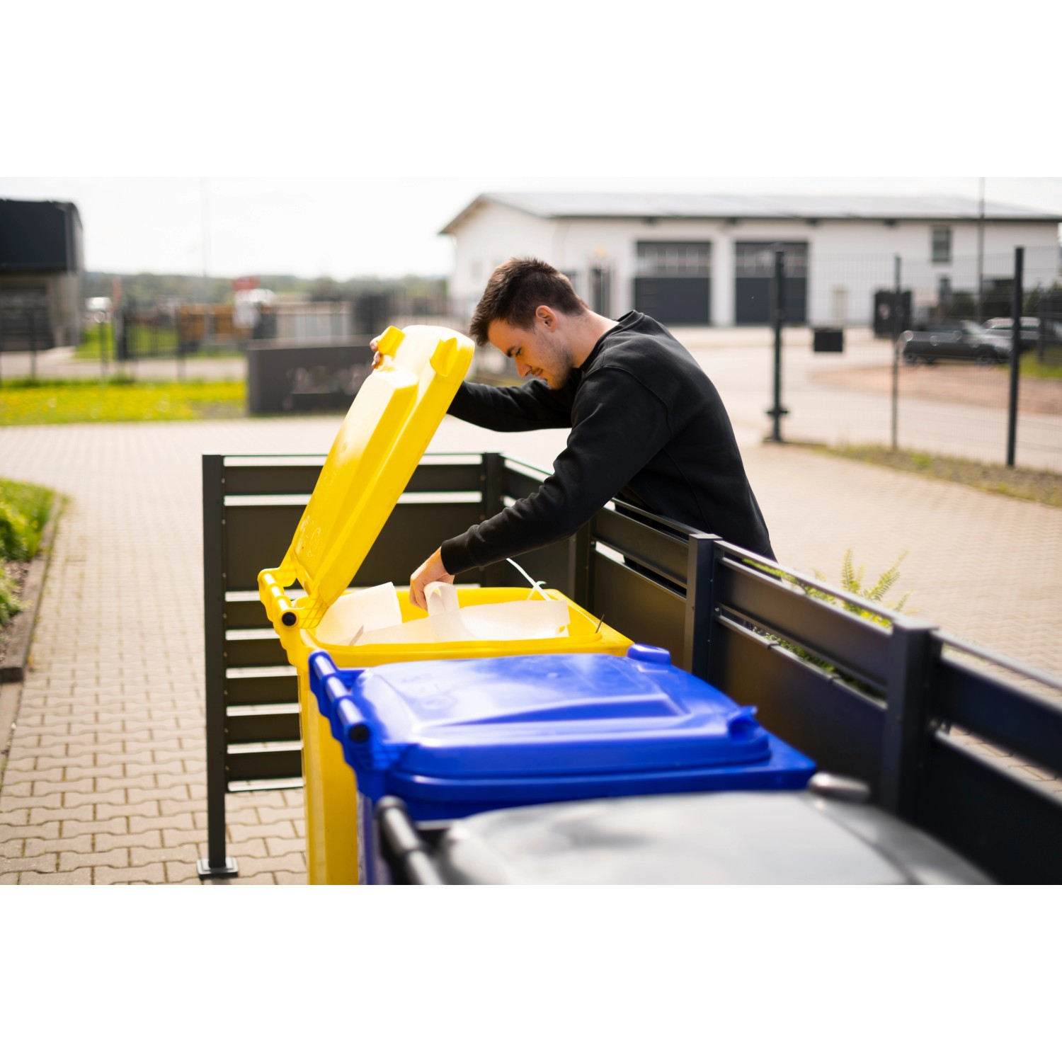 Westmann Mülltonnenverkleidung Planum für 5 Tonnen (je 240 l) in Holzoptik. Mann wirft Müll in gelbe Tonne.