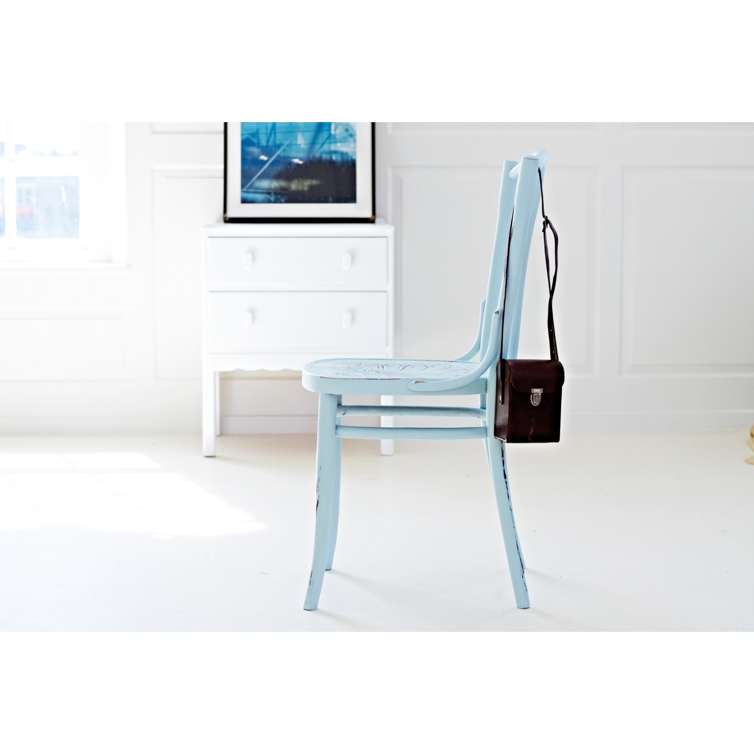 Hellblau lackierter Holzstuhl mit Kreidefarbe und brauner Tasche, im Hintergrund eine Kommode.