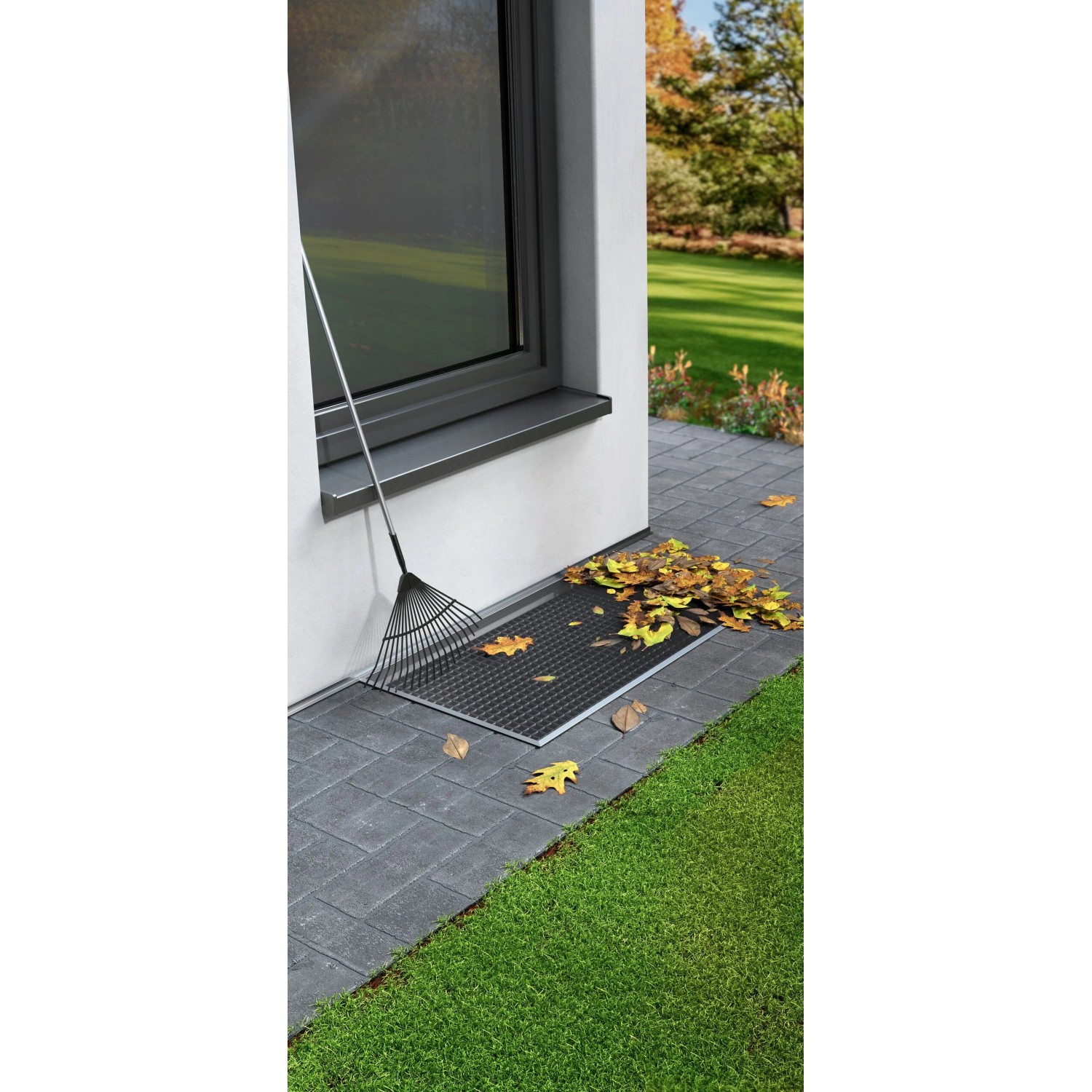 tesa® Insect Stop Lichtschachtabdeckung aus Edelstahl mit Laub und Rechen vor Kellerfenster.