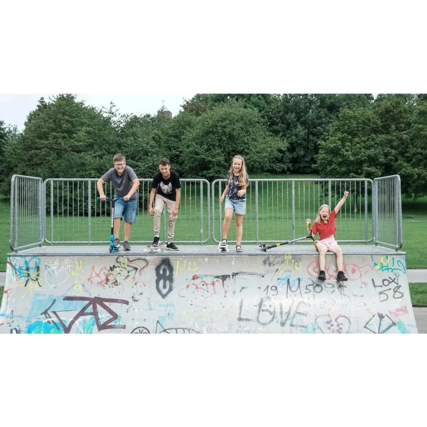 Vier Kinder mit blauem Six Degrees Stuntscooter und Skateboard im Skatepark.