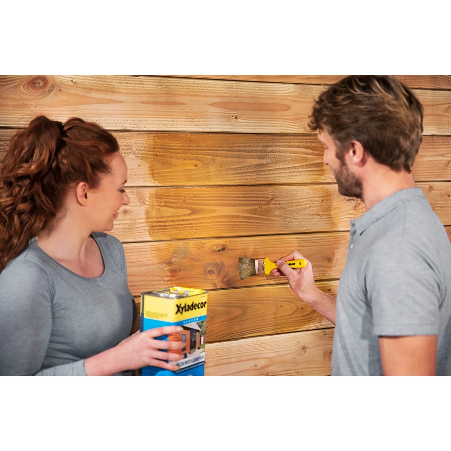 Holzschutz-Lasur Farblos von Xyladecor im Einsatz: Mann streicht Holz, Frau hält Dose.