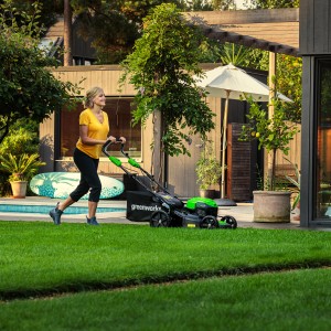 Frau mäht Rasen mit Greenworks Akku-Rasenmäher GD40LM46SP. Der Rasenmäher ist schwarz/grün und hat einen Fangkorb.