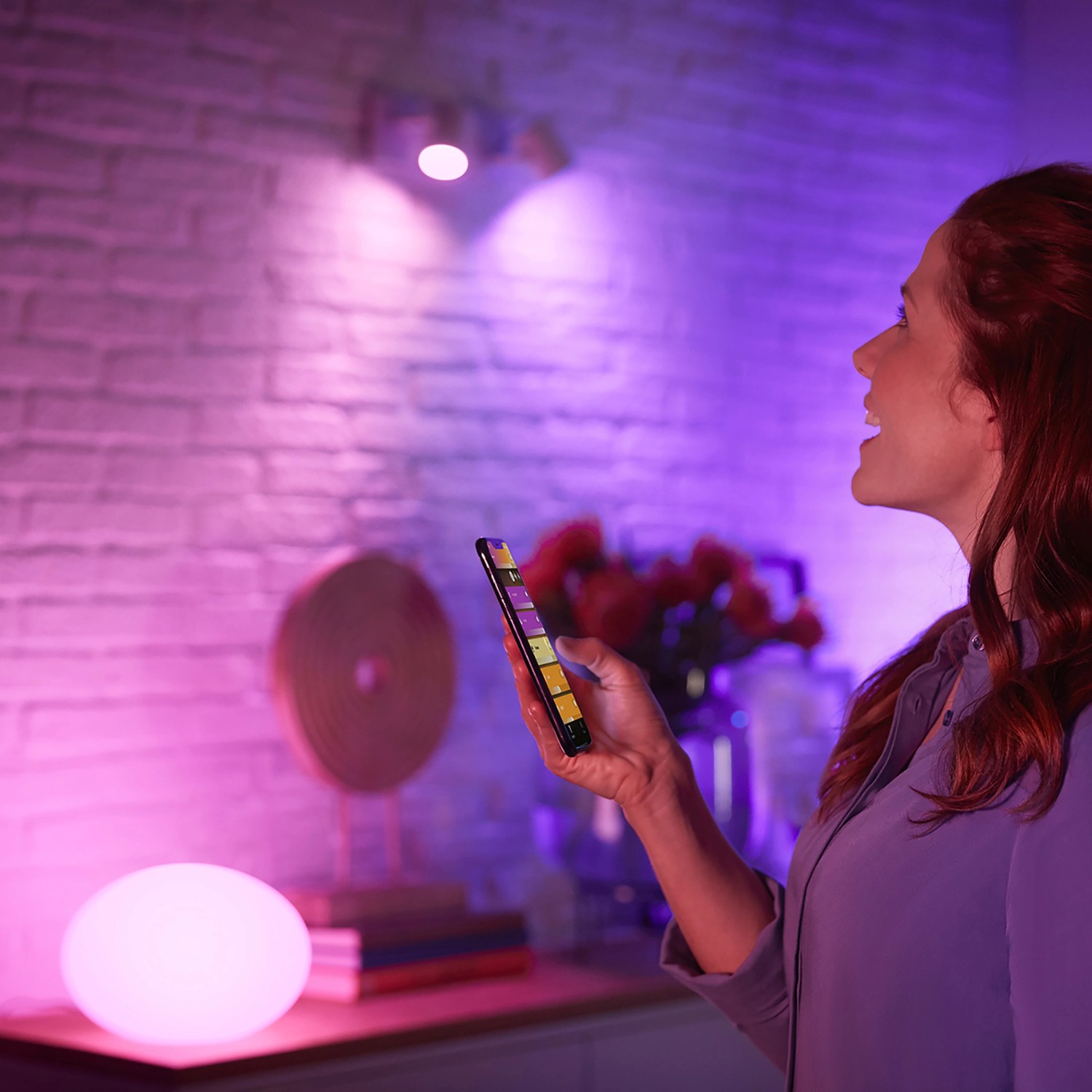 Frau steuert Philips Hue Lampen (2 x E27) per App, farbiges Licht im Raum.