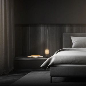 Brilo Tischleuchte Rist Rosegold auf Nachttisch neben Bett in dunklem Schlafzimmer.