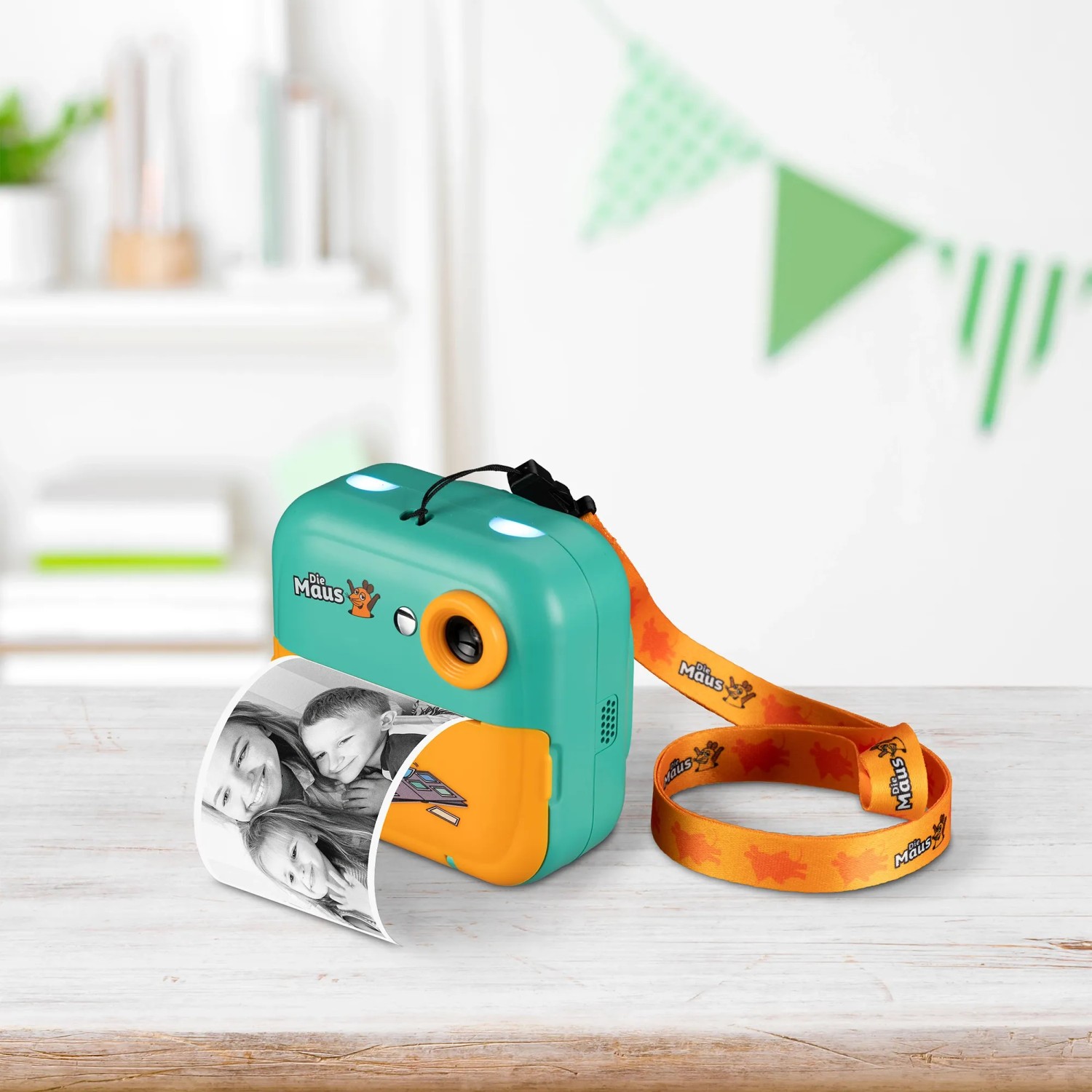 DieMaus Sofortbildkamera für Kinder in Grün-Orange mit Trageriemen und ausgedrucktem Schwarz-Weiß-Foto.