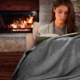 Frau liest gemütlich unter einer hellgrauen Beautissu Alina Kuscheldecke mit Lammfelloptik vor einem Kamin.