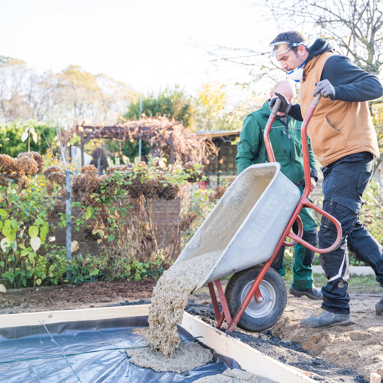 Mann schüttet Pfosten- und Fundamentbeton aus Schubkarre in vorbereitete Fläche.