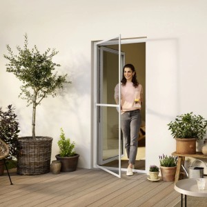 Frau tritt lächelnd durch offenes weißes tesa® Fliegengitter mit Aluminiumrahmen auf Holzterrasse. Insektenschutz für Türen.