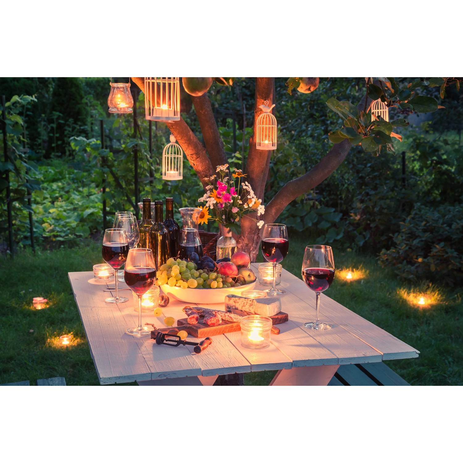 Gartenparty-Szene mit weiß lackiertem Tisch, Wein, Käse und Kerzen im Garten.