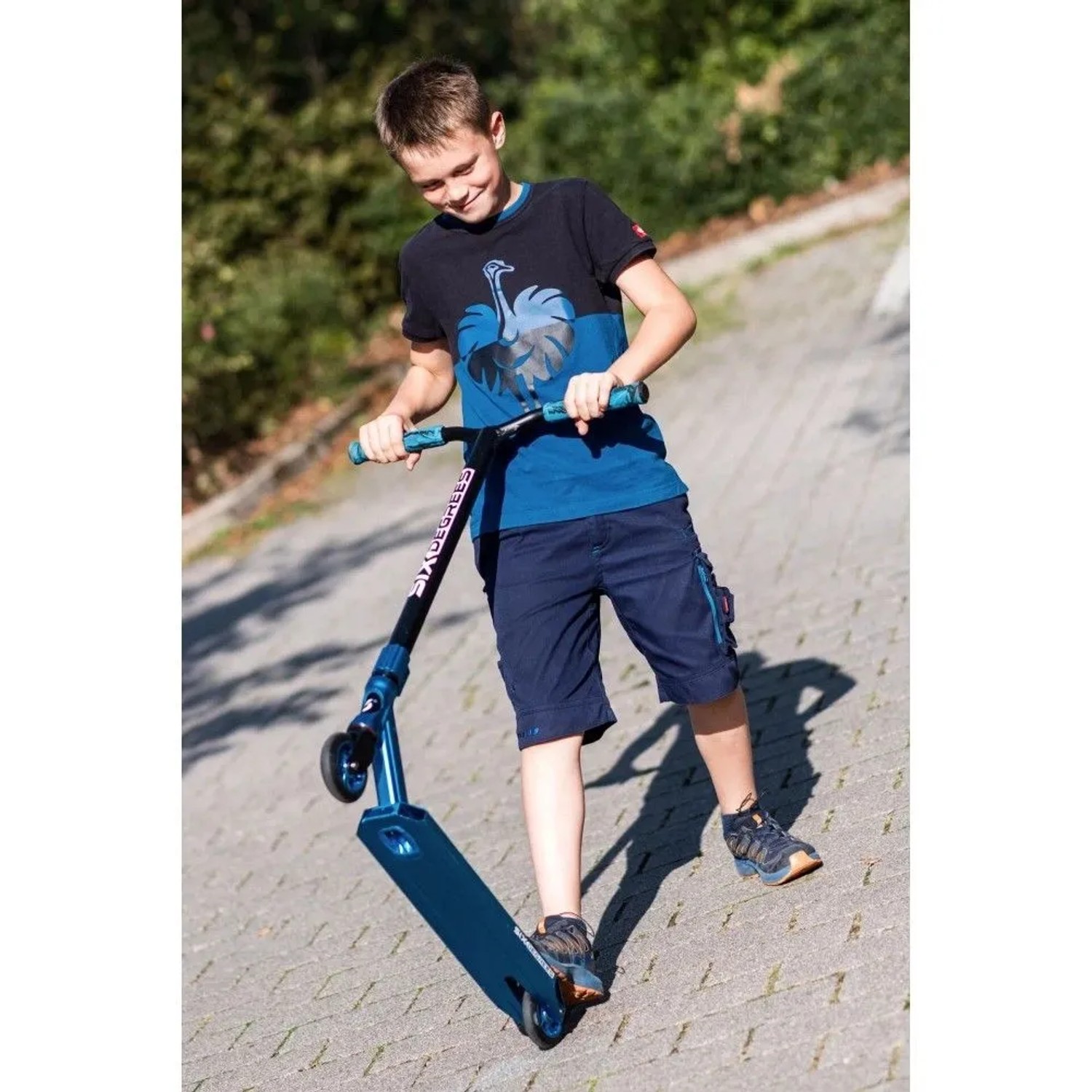 Lachender Junge fährt blauen Six Degrees Stuntscooter mit Wing-Lenker auf gepflastertem Weg. Sport & Fitnessgerät.