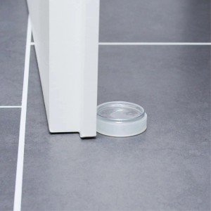 Transparenter Wagner Bodentürstopper Clear (Ø 50 mm) vor weißer Tür auf grauem Fliesenboden.