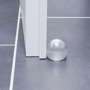 Transparenter, selbstklebender Wagner Bodentürstopper Clear (Ø 45mm) neben einer Tür auf grauem Fliesenboden.