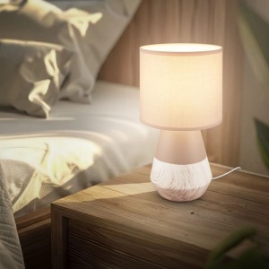 Beige Brilo Tischleuchte Linda mit Holzoptik und Stoffschirm auf Nachttisch.