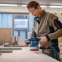 Frau schleift Holz mit blauem Bosch Professional Exzenterschleifer GEX 40-150. Roter Schleifteller, Staubabsaugung, Schutzbrille.