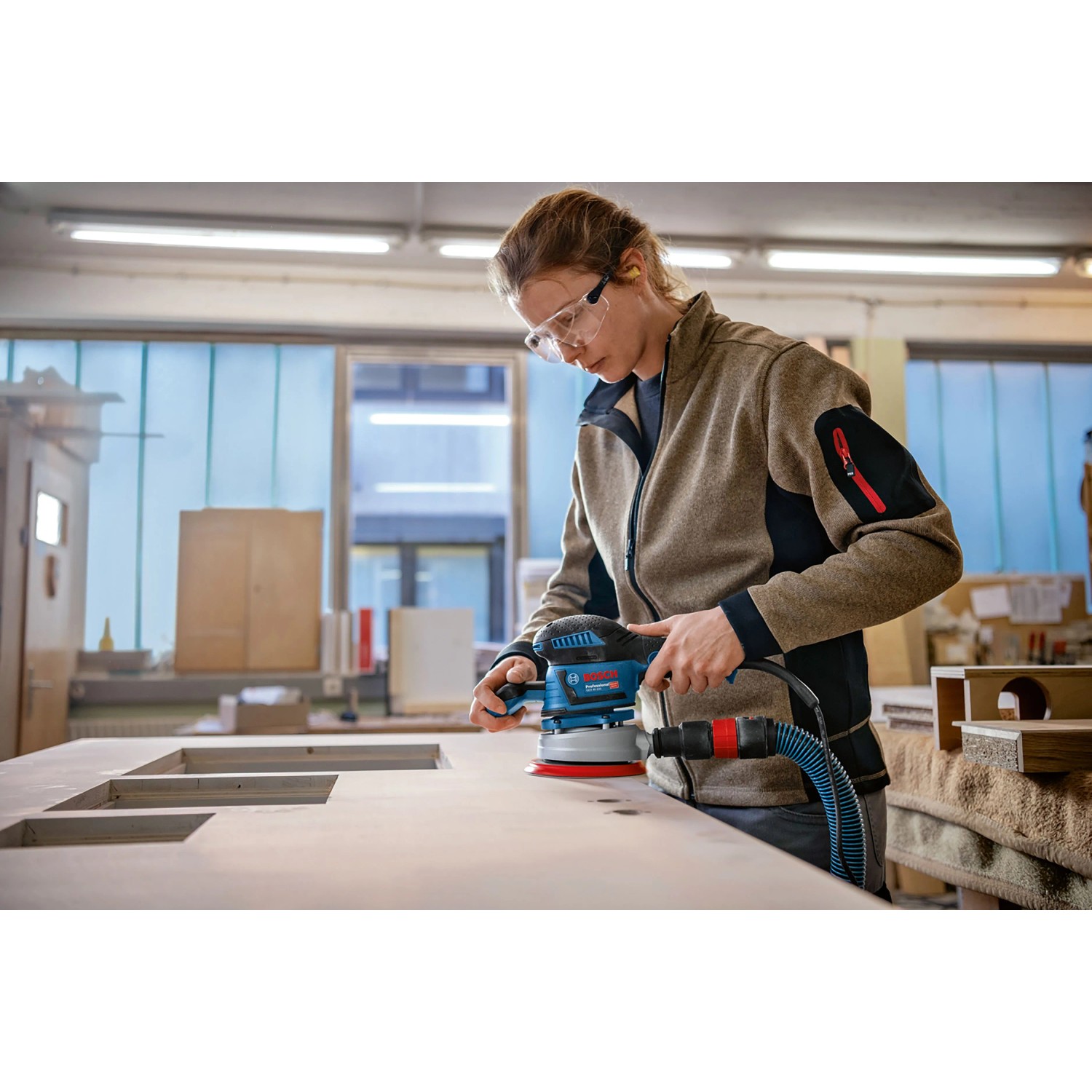 Frau schleift Holz mit blauem Bosch Professional Exzenterschleifer GEX 40-150. Roter Schleifteller, Staubabsaugung, Schutzbrille.