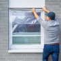 Mann befestigt transparente LUX Fensterschutzfolie am Fensterrahmen.
