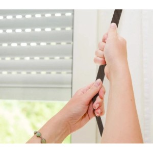Hände ziehen einen braunen Rollladengurt, der einen weißen Rollladen am Fenster bedient. Robuster Gurt für Rollladensteuerung.