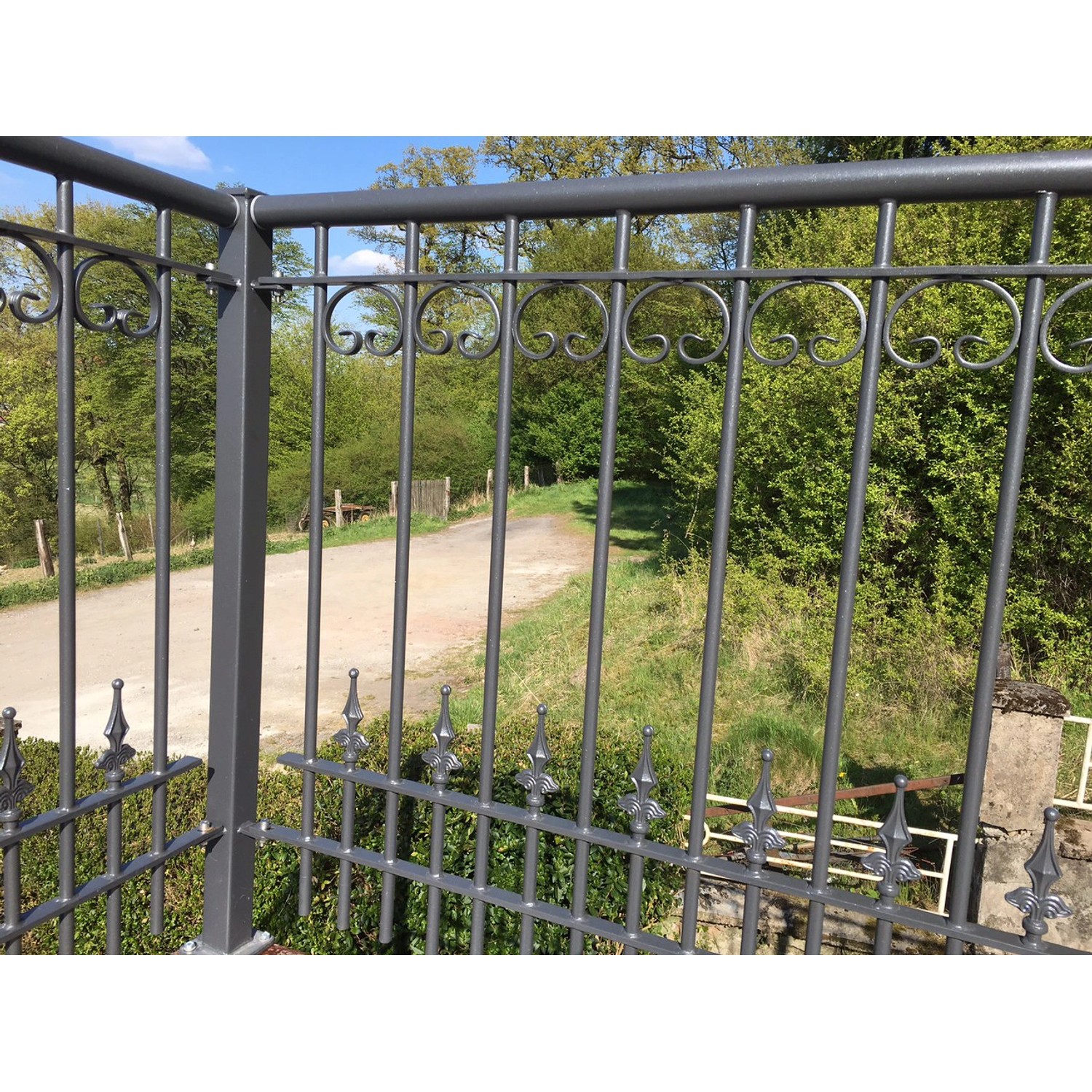 Feuerverzinktes Eleo Geländer Lucca, 96x90 cm, mit Zierspitzen und C-Dekoren vor grüner Landschaft.