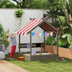 Grauer Holz-Sandkasten mit rot-weiß gestreiftem Dach und Sitzplatz im Garten. Kind spielt mit Sandspielzeug.