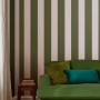 Joules Vliestapete Harborough Stripe Olive Green: Wand mit breiten, grünen und hellen Streifen hinter grünem Sofa.