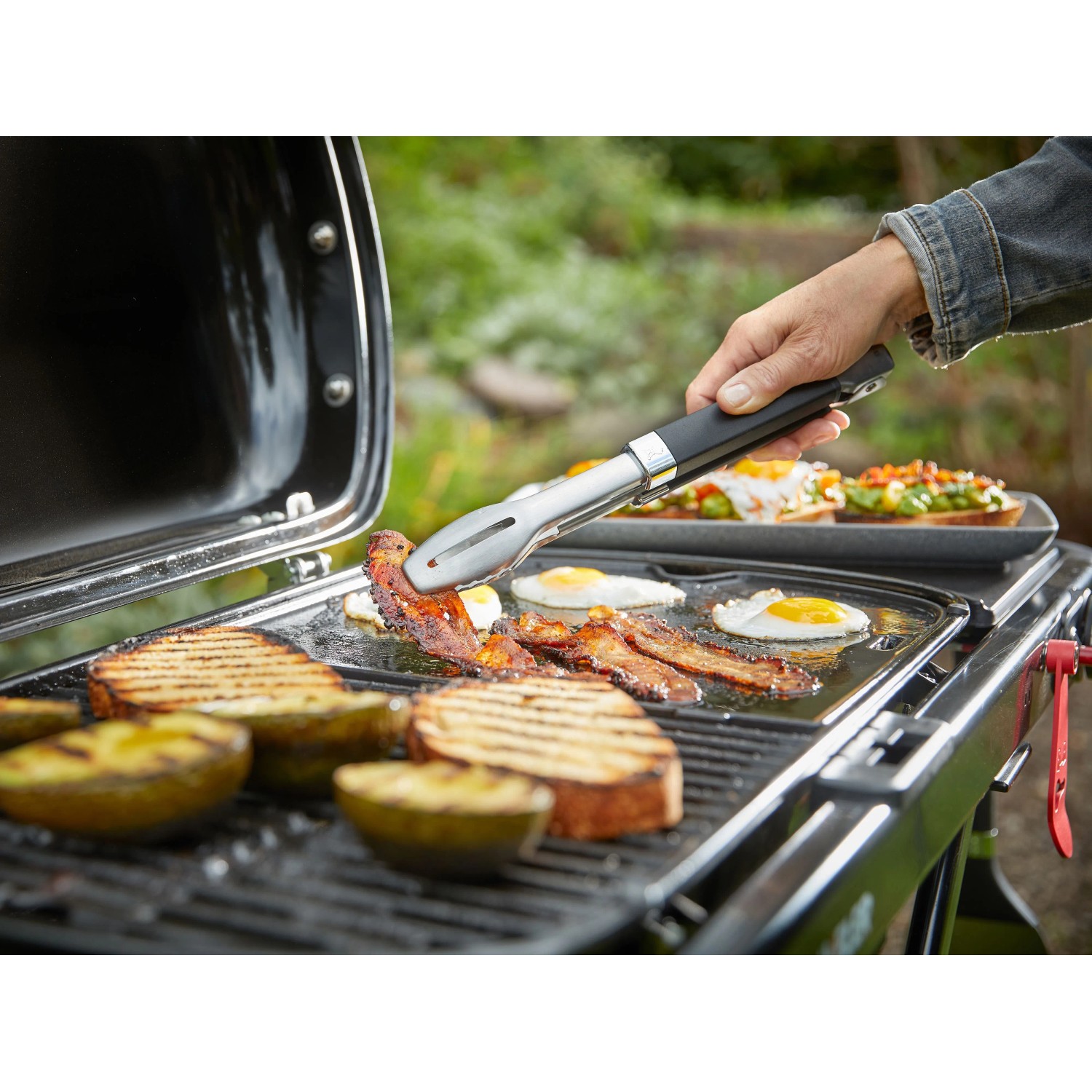Weber Traveler Camping-Gasgrill in Aktion: Grillen von Speck, Eiern und Brot mit Grillzange.