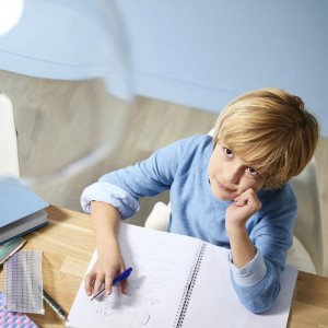 Blonder Junge im blauen Pullover sitzt am Schreibtisch, schreibt in Notizbuch und blickt nachdenklich nach oben.