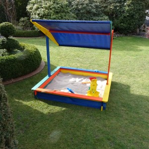 Quadratischer Sandkasten Nadine 140x140 cm mit blauem Sonnensegel im Garten.