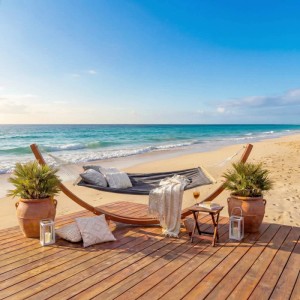 Hängematte mit Holzgestell, braun-graue Matte, Kissen und Beistelltisch auf Holzterrasse am Strand.