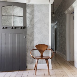 Boutique Vliestapete Royal Palm Moonstone mit Palmenmuster in Grau, kombiniert mit Holzstuhl und grauer Tür.