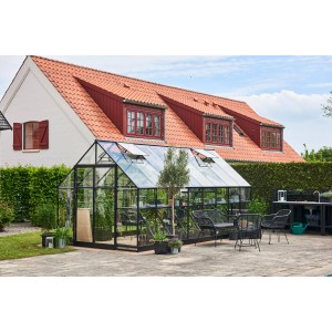 Schwarzes Halls Gewächshaus Qube+ 816 mit 13 m² im Garten, ausgestattet mit Sitzmöbeln.