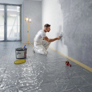 Handwerker trägt Profidur Reibeputz 210 Weiß auf Wand auf; Eimer und Werkzeug im Raum.