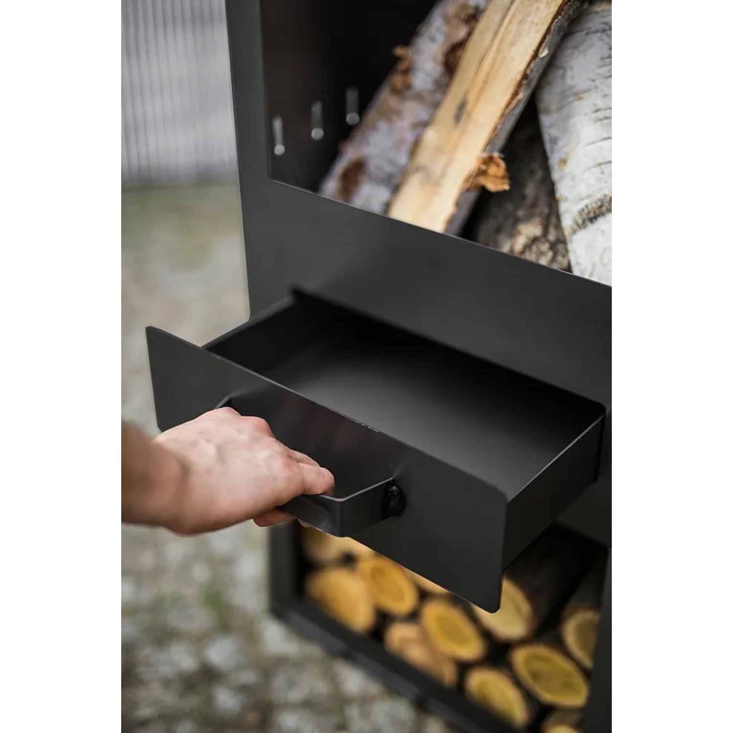 Detailaufnahme: Hand zieht Ascheschublade des CookKing Gartenofens Rosa mit Holzlager.