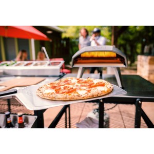 Ooni Pizzaschieber (40 cm) mit Pizza vor Pizzaofen. Grillutensil aus Edelstahl.