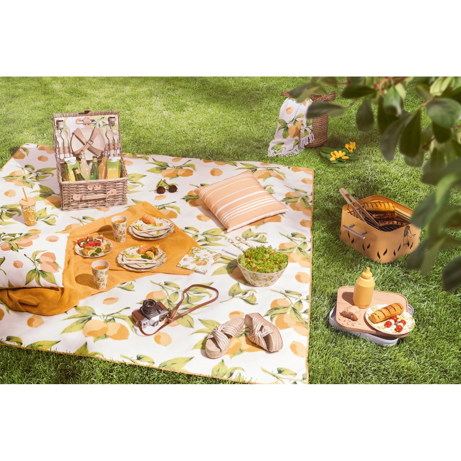 Sommerliches Picknick mit Zitronenmuster: Decke, Kissen, Geschirr, Grill und Getränke im Grünen.