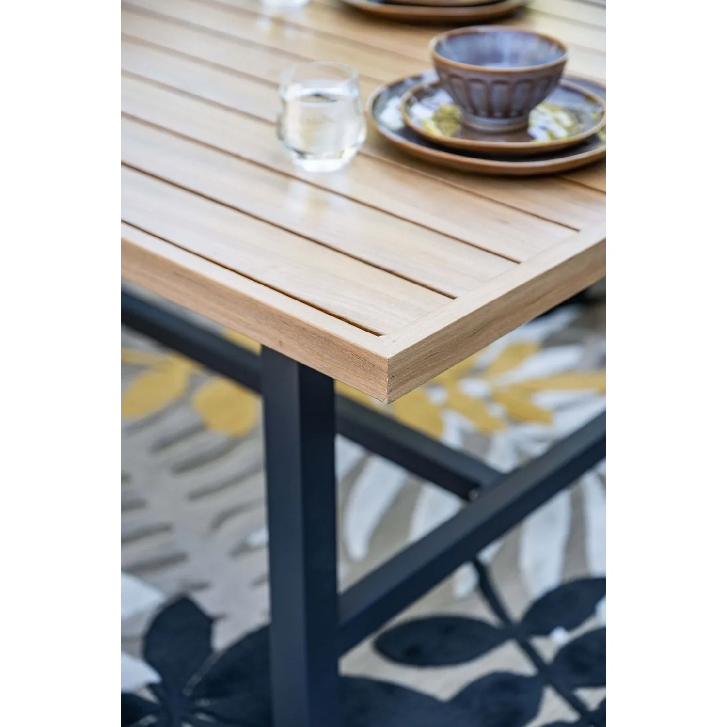 Detail des Elati Gartenmöbel Tisches: Teak-Look-Platte, dunkles Aluminiumgestell, Geschirr auf gemustertem Teppich.
