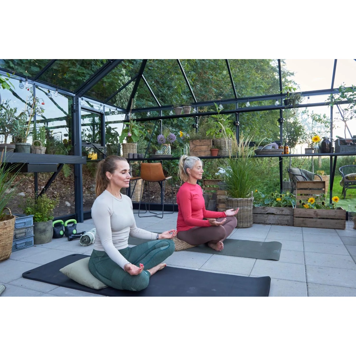 Zwei Frauen machen Yoga im Juliana Gewächshaus Grand Oase 25 Schwarz-Anthrazit, umgeben von Pflanzen.