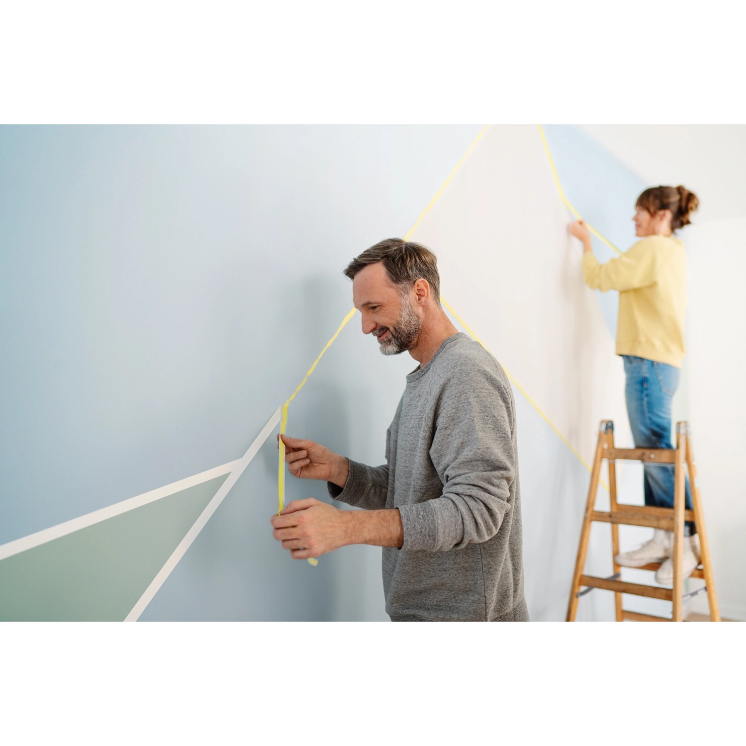 Zwei Personen kleben mit Tesa Malerband eine Wand für farbige geometrische Formen ab.