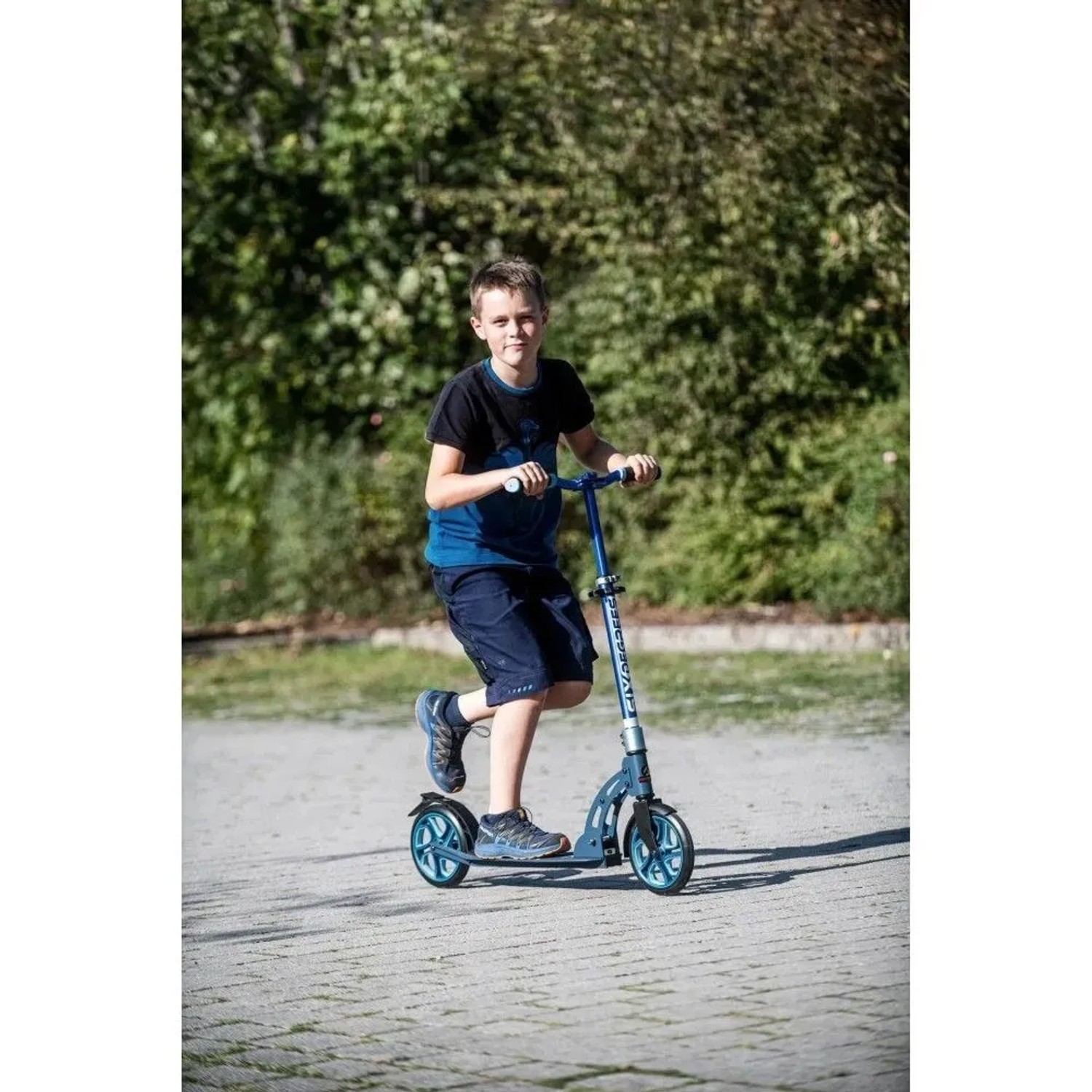 Junge fährt blauen Six Degrees Aluminium Scooter mit 205 mm Rollen auf gepflastertem Weg. Höhenverstellbarer Klappscooter.