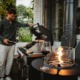 Enders Gas-Terrassenfeuer Nova, schwarz, mit Glaszylinder und Ablage, im Hintergrund ein Grill.