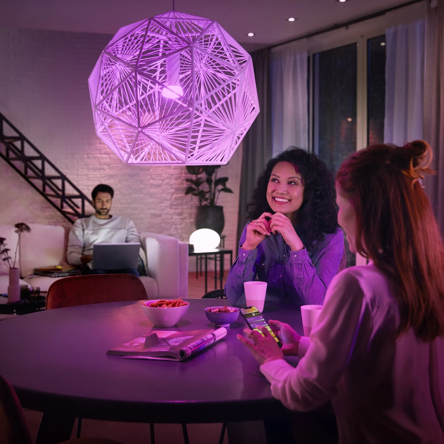 Szene mit Philips Hue Lampe und Bridge: Zwei Frauen am Tisch, Mann mit Laptop im Hintergrund.