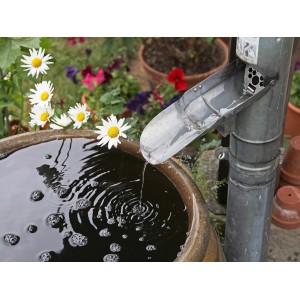 Regenrohrklappe NW 60 aus Aluminium leitet Wasser in ein Becken, umgeben von Blumen.