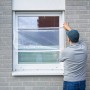 Mann befestigt transparente LUX Fensterschutzfolie am Fensterrahmen.