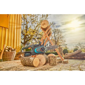 Gardena Akku-Kettensäge PowerSaw 250/18V Solo auf Holzstamm, im Hintergrund Holzstapel und Baum.