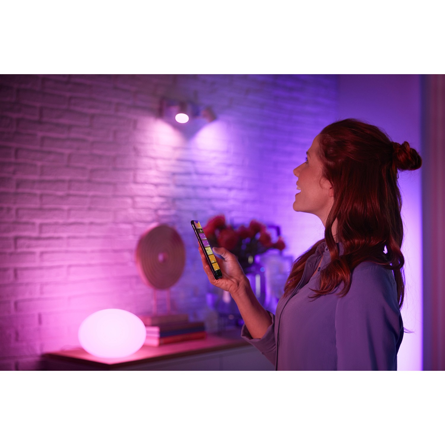 Frau steuert Philips Hue E27 LED Lampe mit Smartphone, farbige Beleuchtung im Raum.