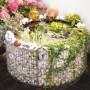 Runder Gabionen-Hochteich (Ø 92cm) mit Steinen, Pflanzen und Deko-Elementen im Garten.