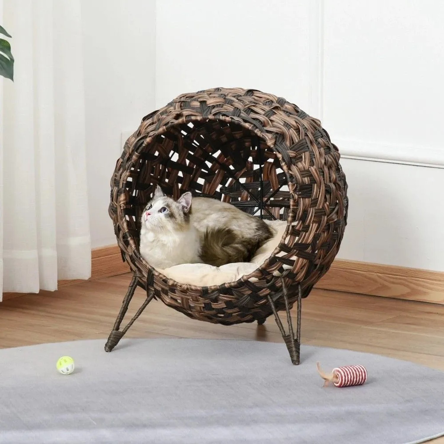 Rundes PawHut Katzenhaus aus dunklem Rattan mit weichem Kissen, Katze liegt darin. Erhöhte Katzenhöhle auf Beinen.