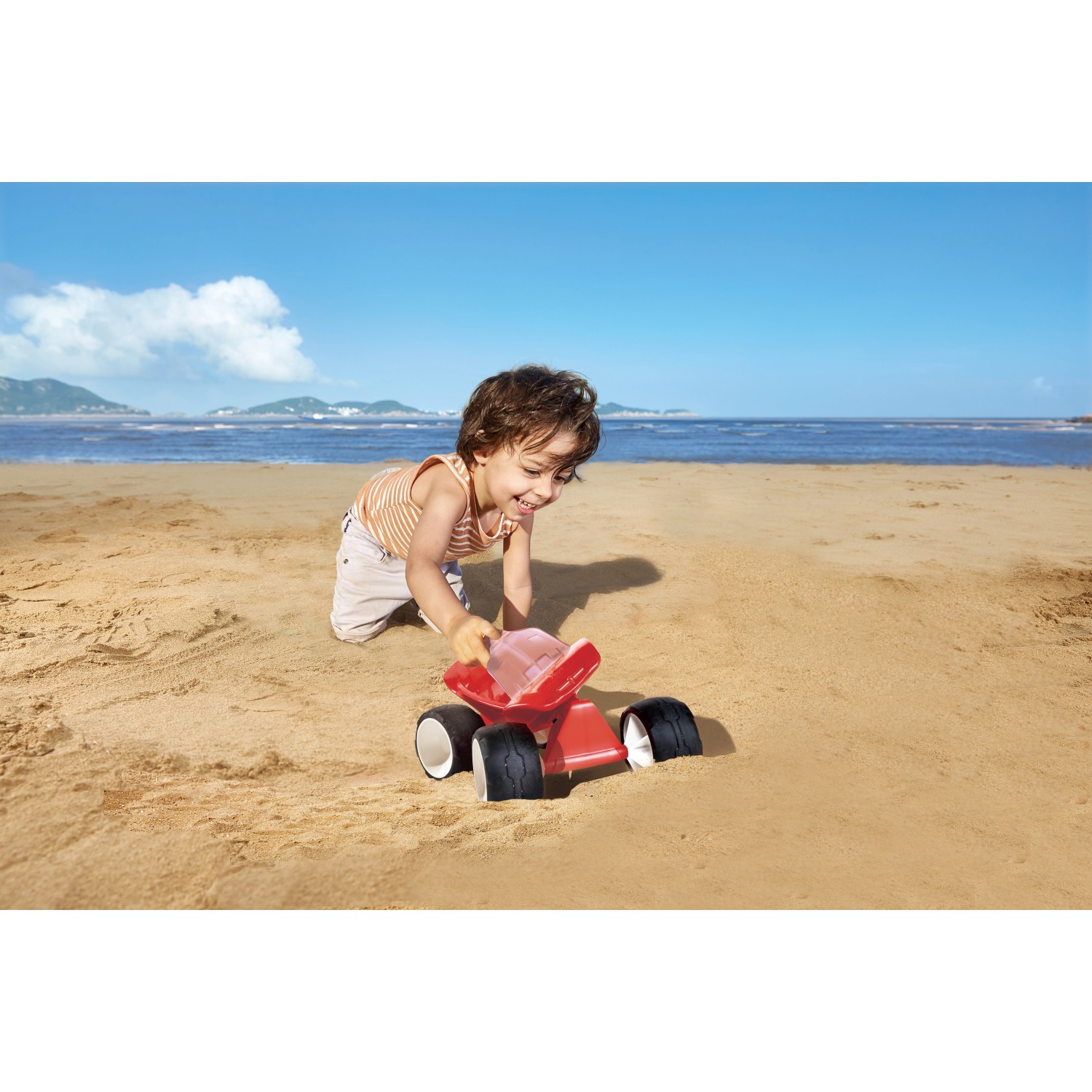 Kind spielt mit rotem Hape Dünen-Buggy Sandspielzeug am Strand.