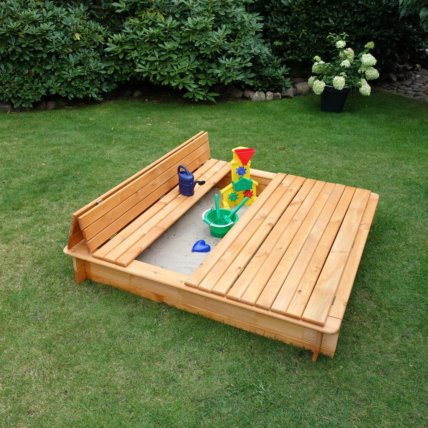 Holz Sandkasten Donald 140x140 cm mit klappbarer Abdeckung und Spielzeug im Garten.