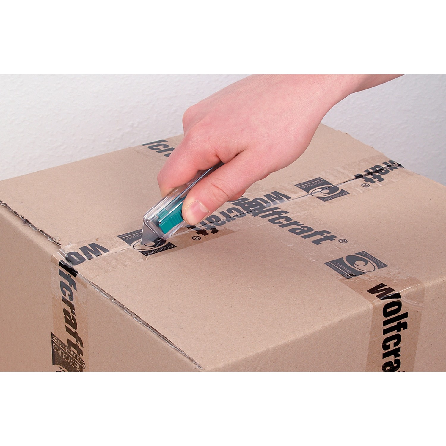 Hand schneidet Paket mit wolfcraft Sicherheits-Trapezklingen-Messer. Kunststoffgehäuse.