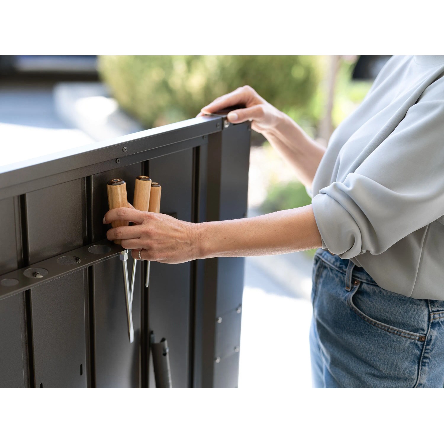 Person verstaut Gartenwerkzeug in Halterung der dunkelgrauen Biohort Gartenbox HighBoard. Praktische Aufbewahrung für Universalbox.
